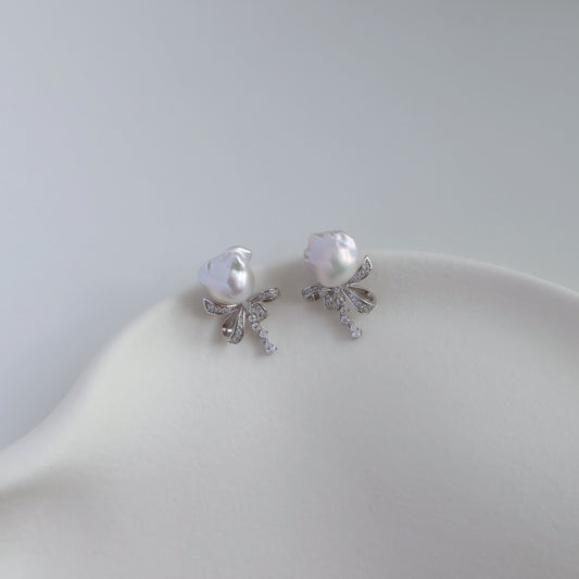 A pair of silver-toned stud earrings featuring a baroque pearl and a small bow design accented with clear crystals, displayed on a white jewelry plate against a light gray background.