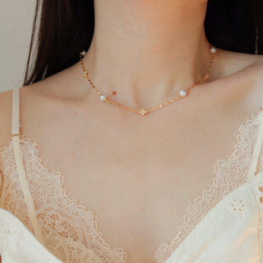 A delicate gold chain necklace with small white pearl beads, worn on a model's neck over a cream lace camisole.