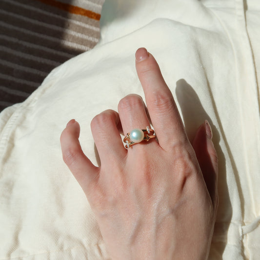 A close-up of a hand wearing a gold ring with a central white pearl, resting on a cream-colored fabric surface.