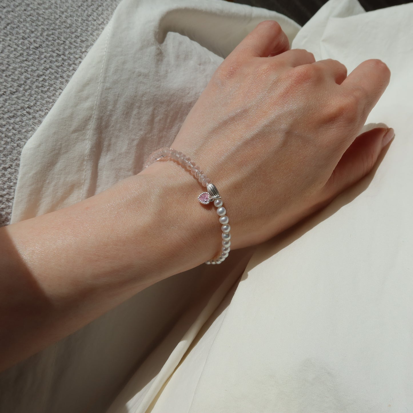 Hand wearing a bracelet with a pink heart charm on a neutral background