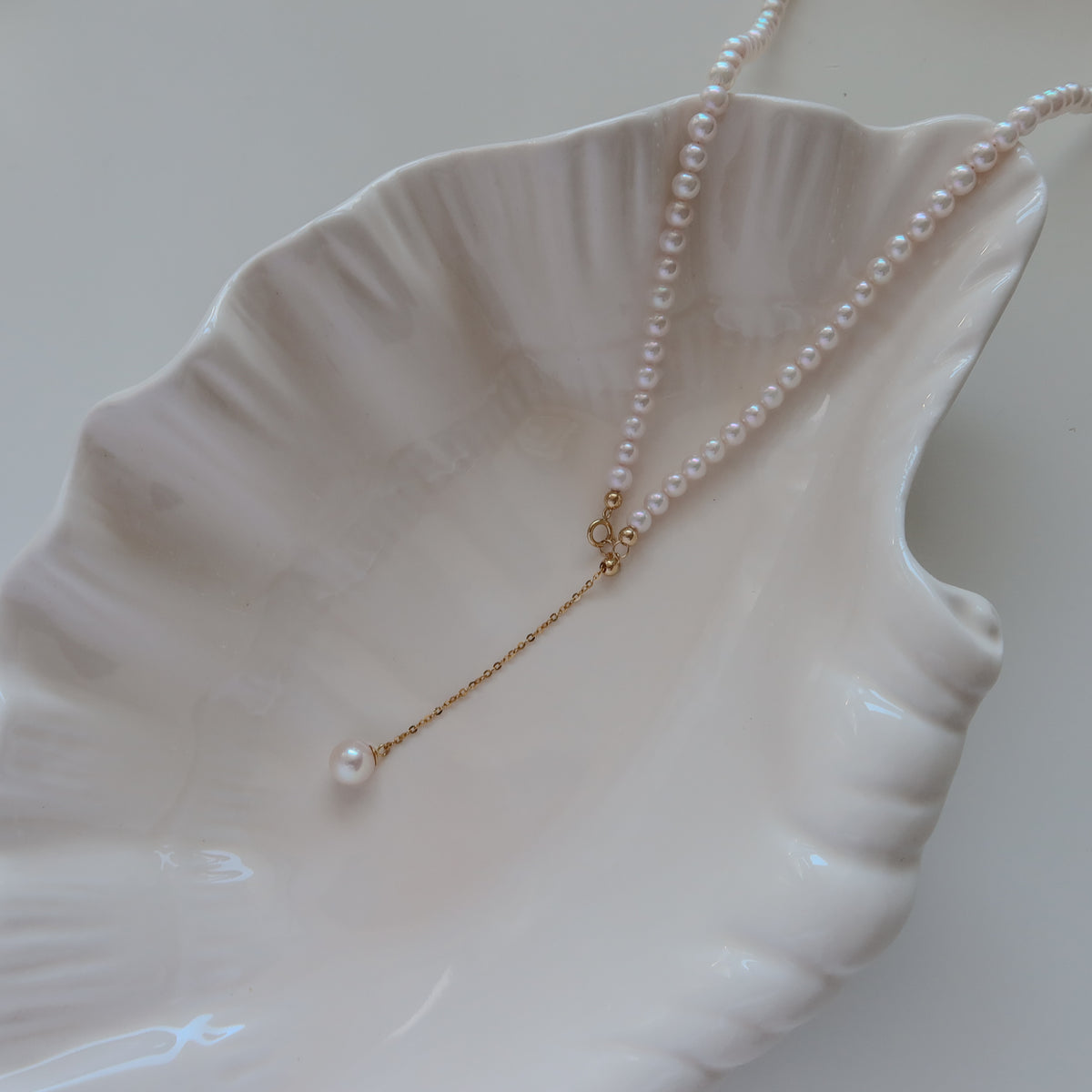 18k Gold Akoya Pearl Necklace resting on a white ceramic shell dish, highlighting its lustrous white Akoya pearls and delicate chain drop detail.
