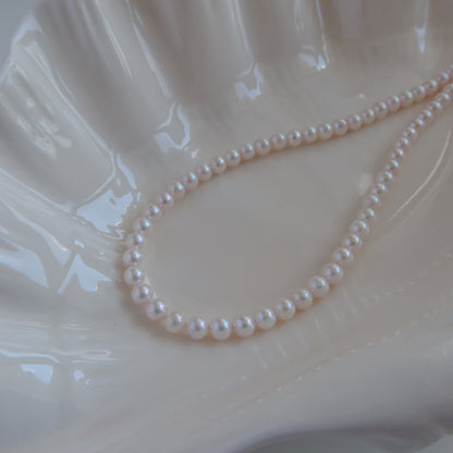 18k Gold Akoya Pearl Necklace resting on a white ceramic shell dish, showcasing its lustrous white Akoya pearls and delicate chain drop detail.