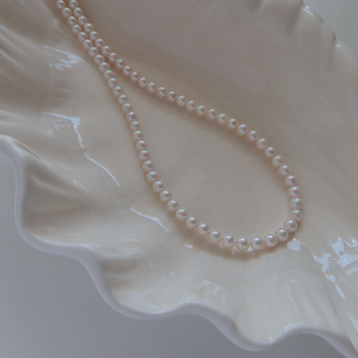 18k Gold Akoya Pearl Necklace resting on a white ceramic shell dish, showcasing its lustrous white Akoya pearls and delicate chain detail.