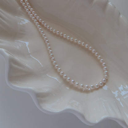 18k Gold Akoya Pearl Necklace resting on a white ceramic shell dish, showcasing its lustrous white Akoya pearls and delicate chain detail.