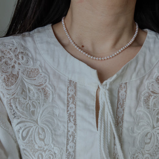 Close-up of a person wearing the 18k Gold Akoya Pearl Necklace, highlighting its lustrous white Akoya pearls and delicate chain drop detail against a white lace top.