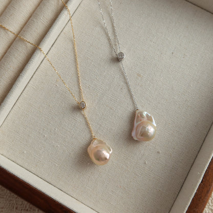 Two delicate pendant necklaces displayed on a beige jewelry box. One necklace is gold-toned baroque pearl with a small round cz stone and a large irregular pearl pendant.