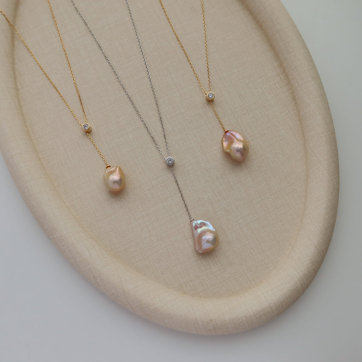 Three delicate necklaces with baroque pearl pendants and small CZ accents, displayed on a beige fabric jewelry stand.