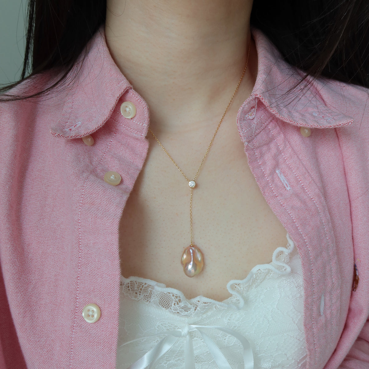 A close-up of a person wearing a delicate gold lariat necklace with a baroque pearl pendant, styled over a pink button-up shirt and a white lace top.