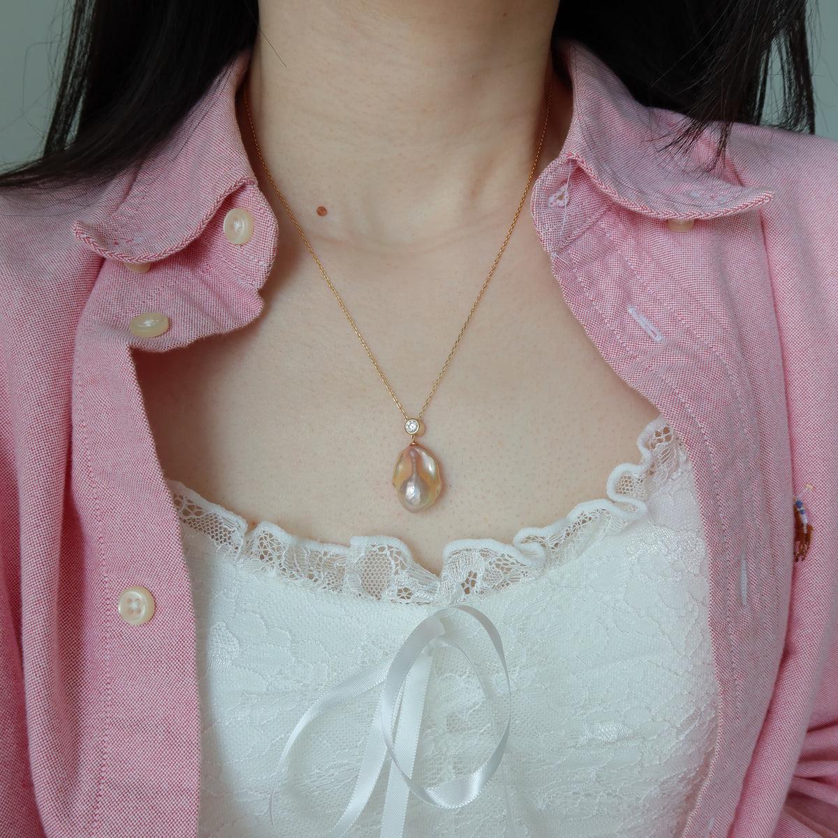 A close-up of a person wearing a delicate gold chain necklace with a pink baroque pearl pendant, styled over a white lace camisole and a pink button-up shirt.