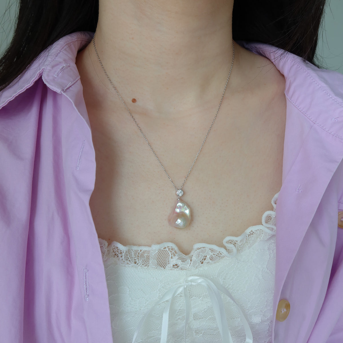A close-up of a person wearing a delicate silver-toned necklace with a light purple baroque pearl pendant, styled with a light purple button-up shirt and a white lace top.