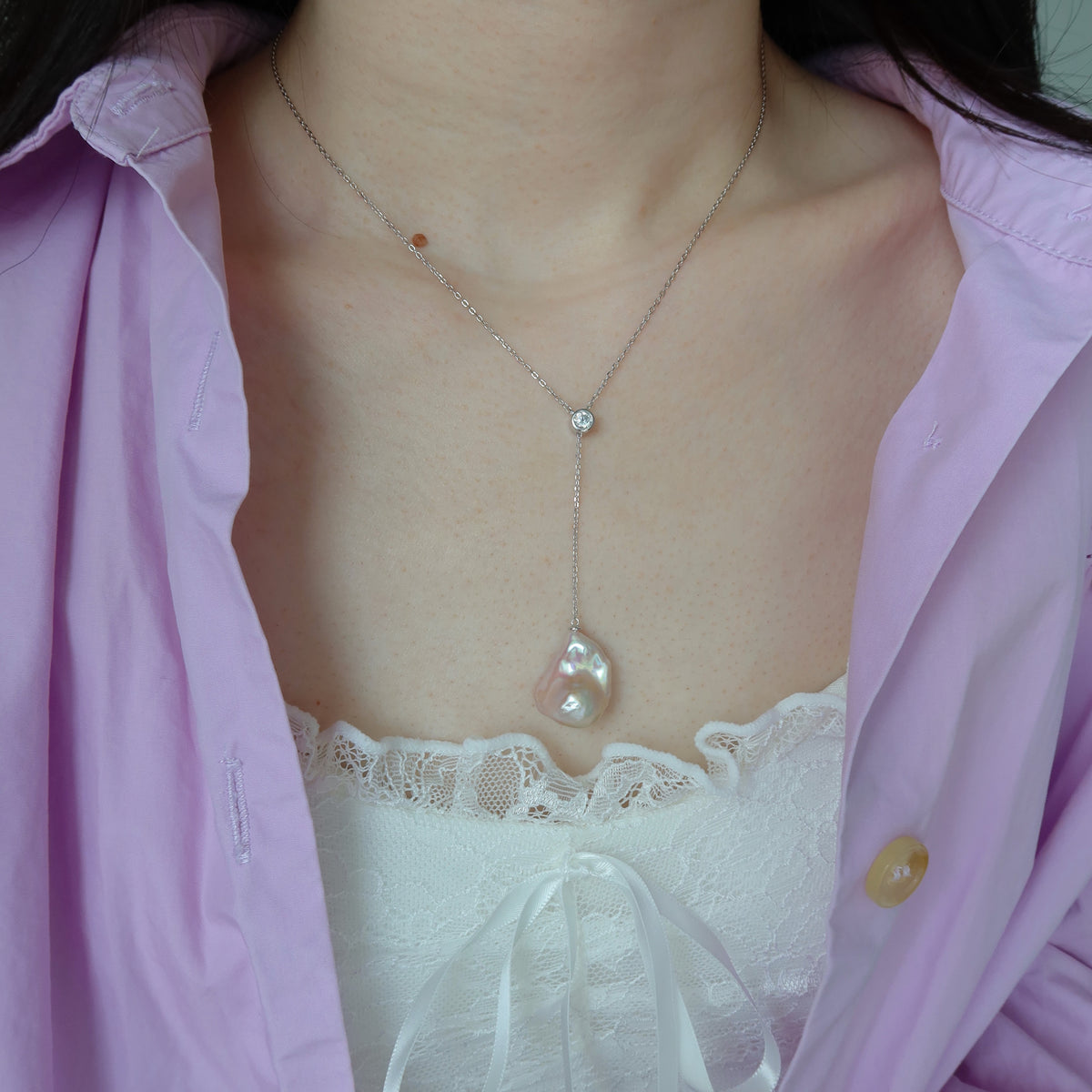 A close-up of a person wearing a delicate silver Y-necklace with a large irregular pearl pendant and a small clear stone at the collarbone, styled with a lavender shirt and white lace top.