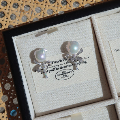 Coquette Baroque Pearl Earrings displayed inside a presentation box on a vintage-style card, highlighting their white baroque pearls and sparkling pavé ribbon bow studs.