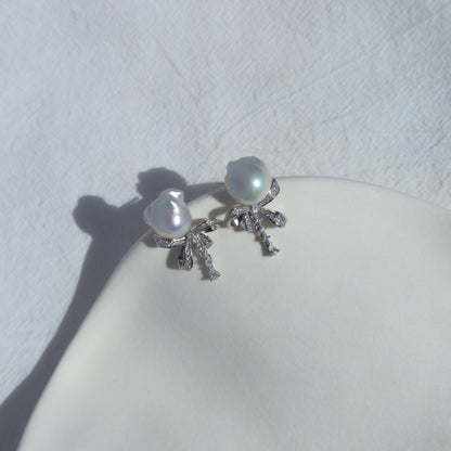 Coquette Baroque Pearl Earrings resting on a white ceramic dish in natural sunlight, highlighting their luminous white baroque pearls and sparkling pavé ribbon bow studs.