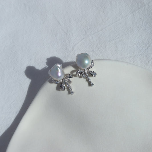 Coquette Baroque Pearl Earrings resting on a white ceramic dish in natural sunlight, highlighting their luminous white baroque pearls and sparkling pavé ribbon bow studs.