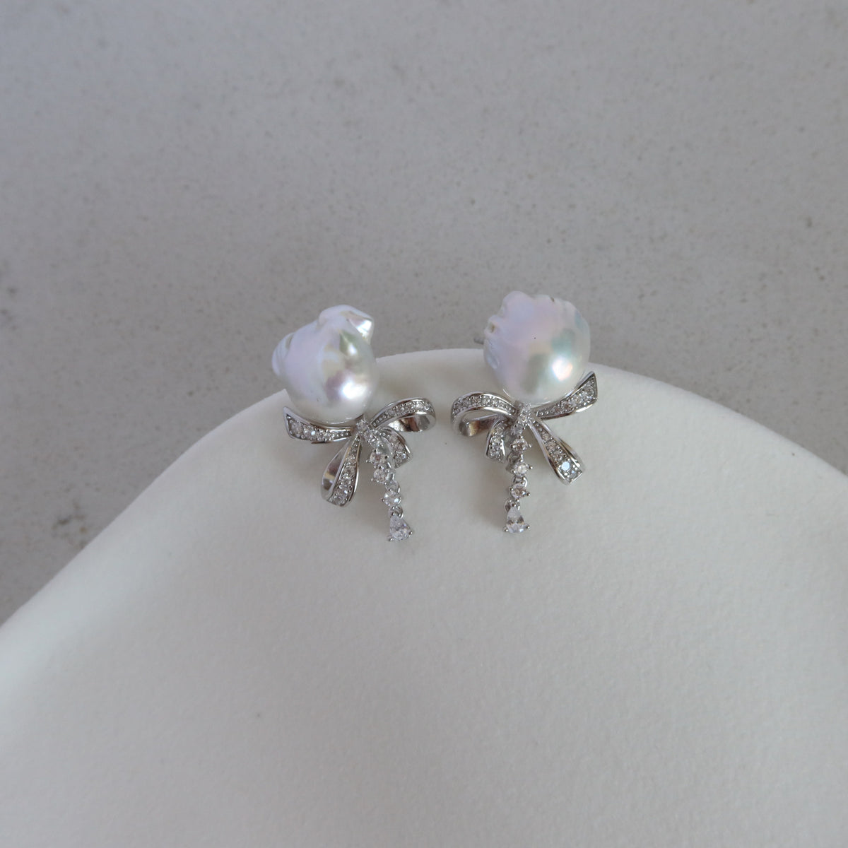 Coquette Baroque Pearl Earrings displayed on a white jewelry stand, showcasing their iridescent white baroque pearls and sparkling pavé ribbon bow studs with dangling stone accents.