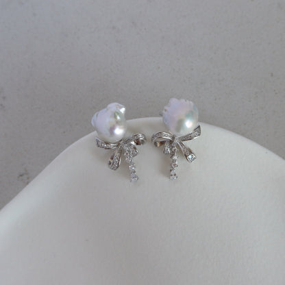 Coquette Baroque Pearl Earrings displayed on a white jewelry stand, showcasing their iridescent white baroque pearls and sparkling pavé ribbon bow studs with dangling stone accents.