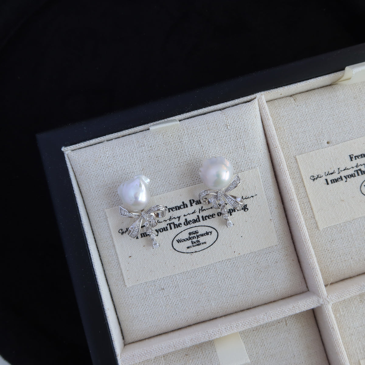 Coquette Baroque Pearl Earrings displayed inside a presentation box on a cream-colored insert with French script, showcasing their white baroque pearls and sparkling pavé ribbon bow studs.