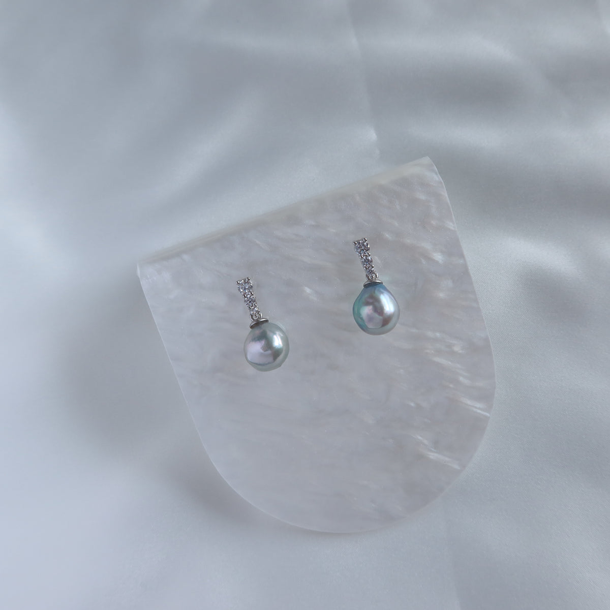 Close-up of Adeline Blue Akoya Baroque Pearl Earrings resting on a white patterned dish against a soft fabric background, showcasing their iridescent blue akoya baroque pearls and pavé stone settings.