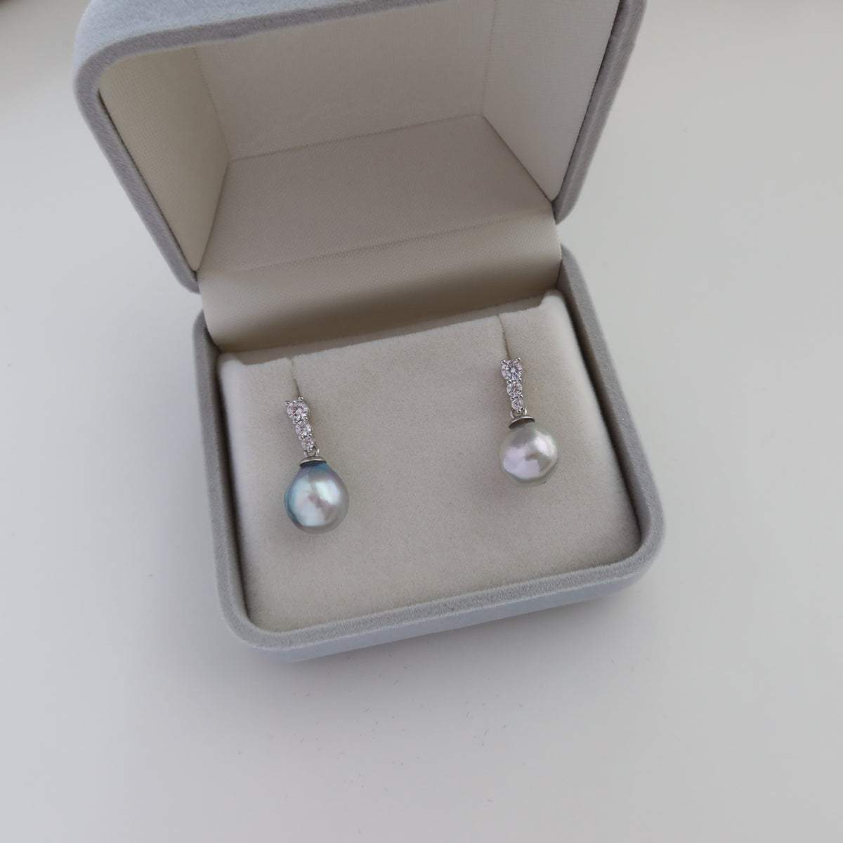 Adeline Blue Akoya Baroque Pearl Earrings displayed inside a gray velvet presentation box, highlighting their iridescent blue akoya baroque pearls and sparkling pavé stone settings.