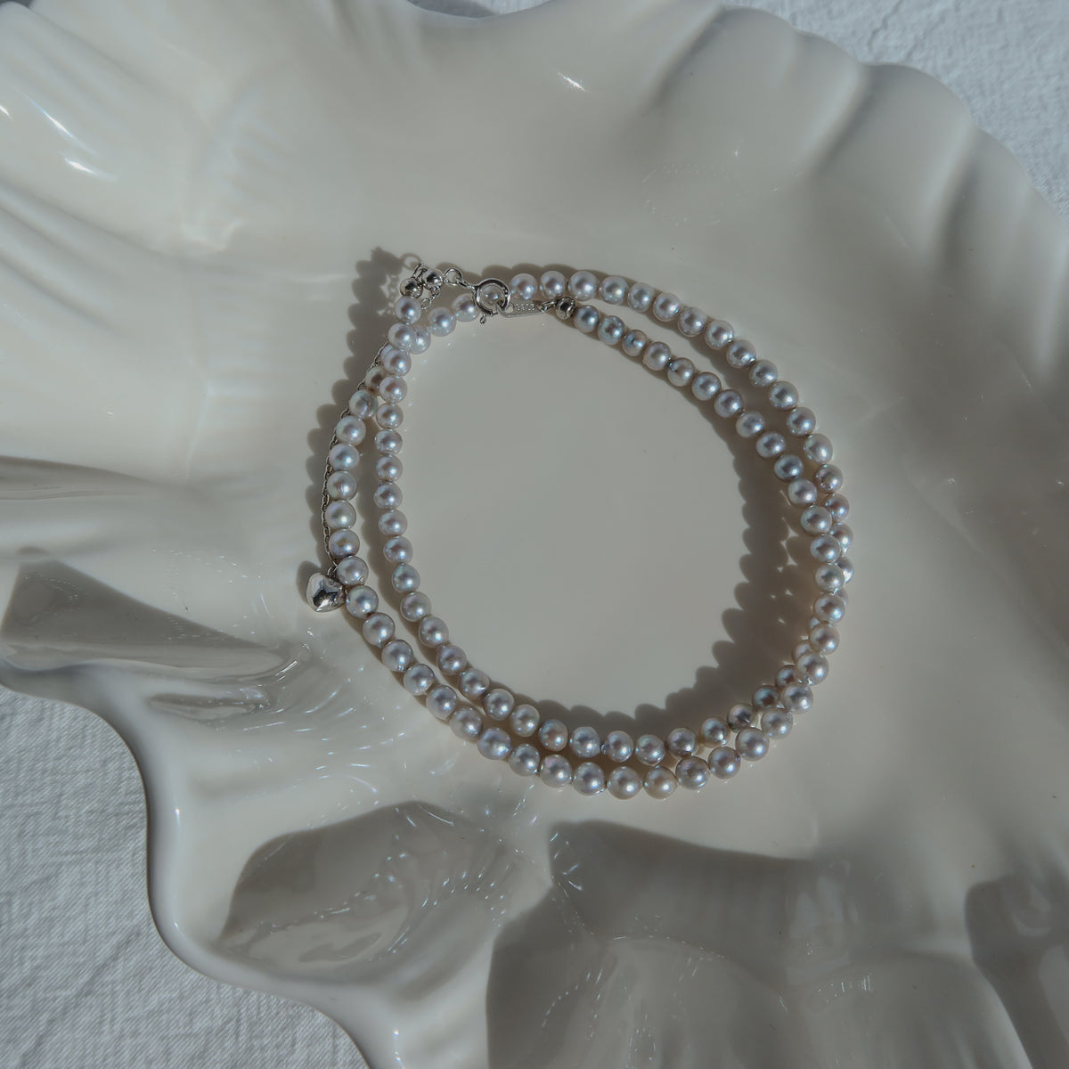 Blue Akoya Pearl Necklace coiled on a white ceramic shell dish in soft natural light, highlighting its lustrous blue Akoya pearls and delicate chain detail.