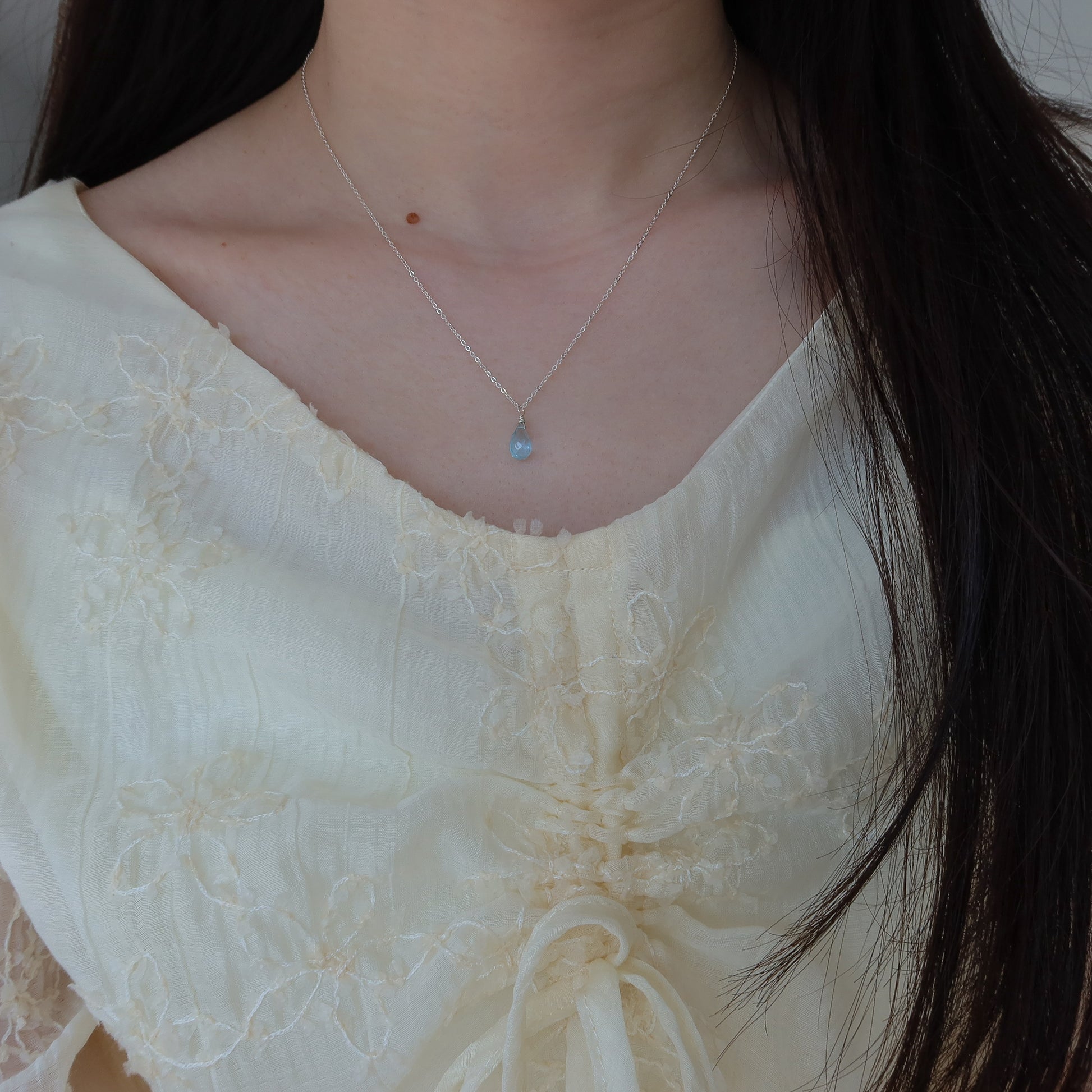 Close-up of a person wearing a delicate blue topaz necklace with a light-colored top.