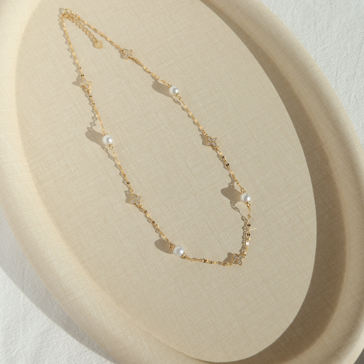 A delicate gold chain necklace with small clover charms and white pearl beads, displayed on a beige textured oval tray.