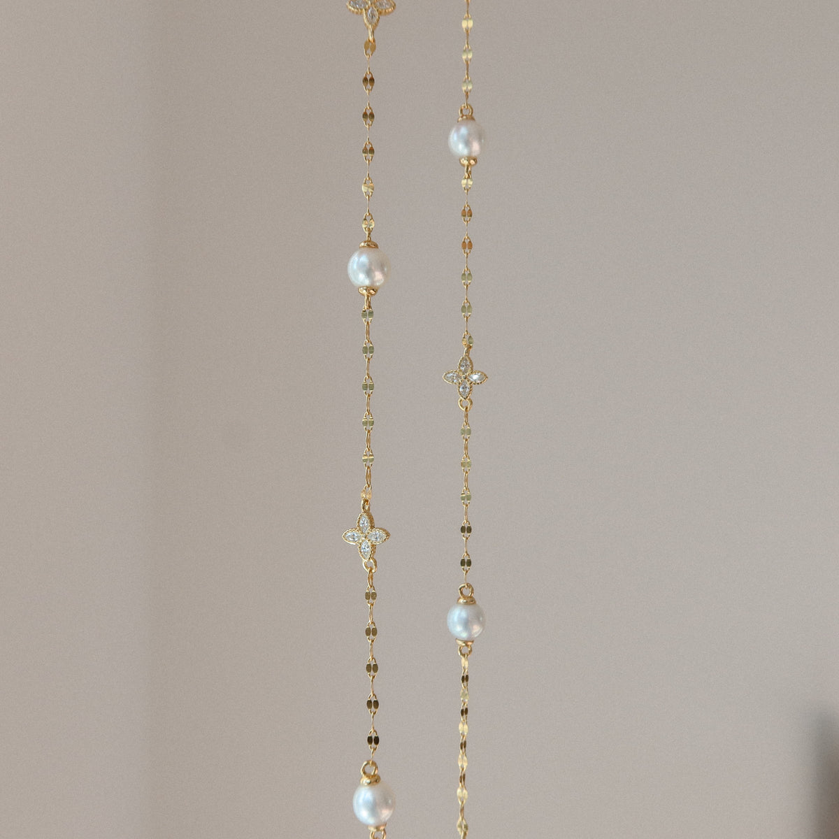 A delicate gold petal chain necklace featuring white pearl beads and small gold clover motifs, hanging against a soft beige background.