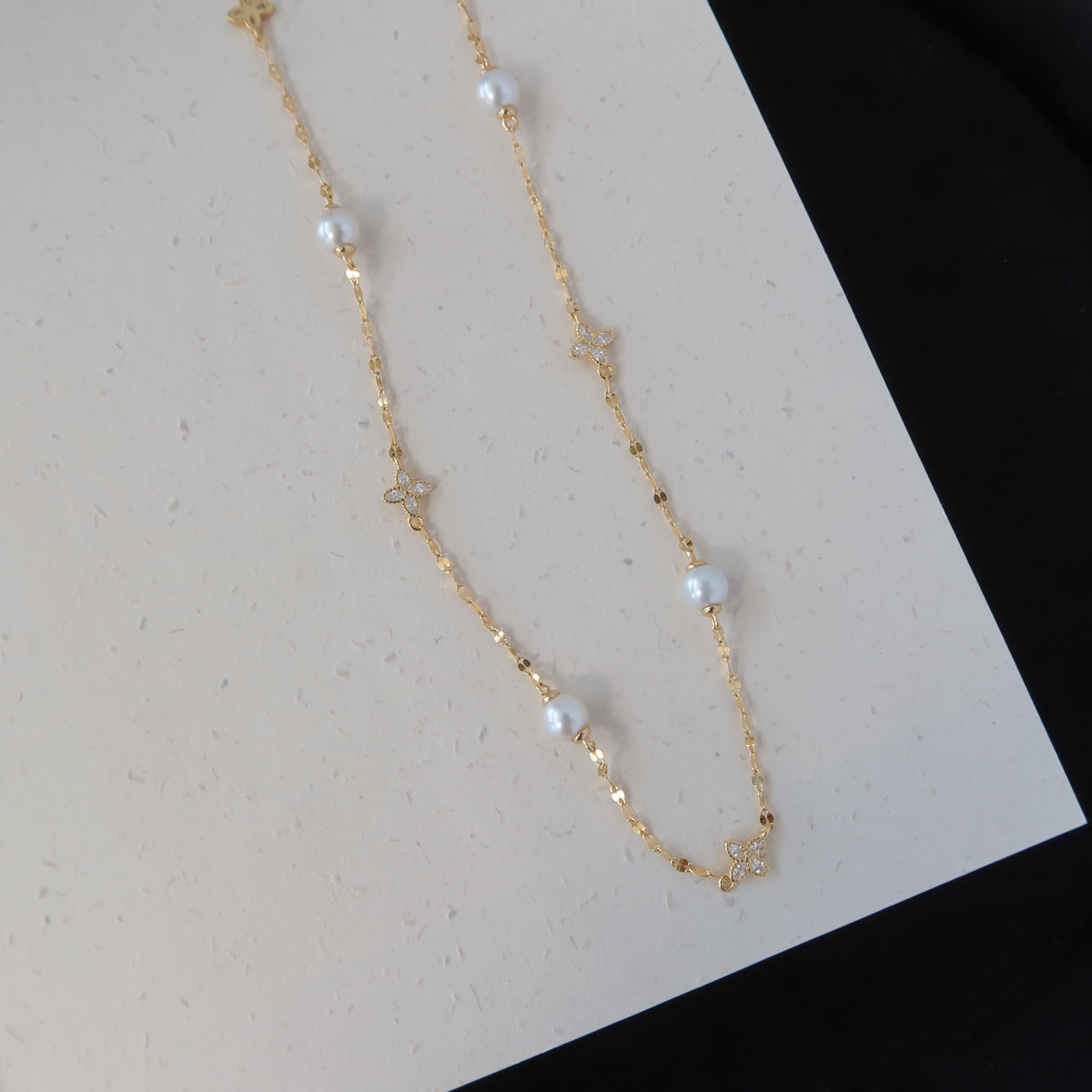 A delicate gold chain necklace featuring small white pearl beads and tiny gold star charms, laid out on a white textured surface with a black background visible in the corner.