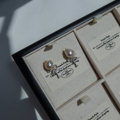 Coquette Baroque Pearl Earrings resting on a canvas jewelry tray, showcasing their pink-toned baroque pearls and sparkling pavé ribbon bows.