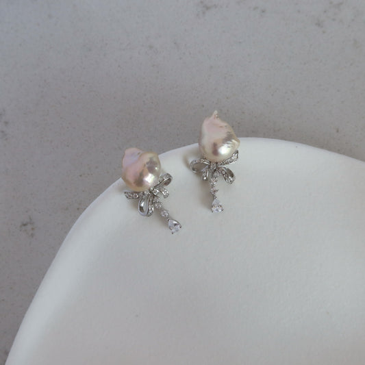 Close-up of Coquette Baroque Pearl Earrings resting on a white ceramic dish, showcasing their pink-toned baroque pearls and pavé ribbon bows.