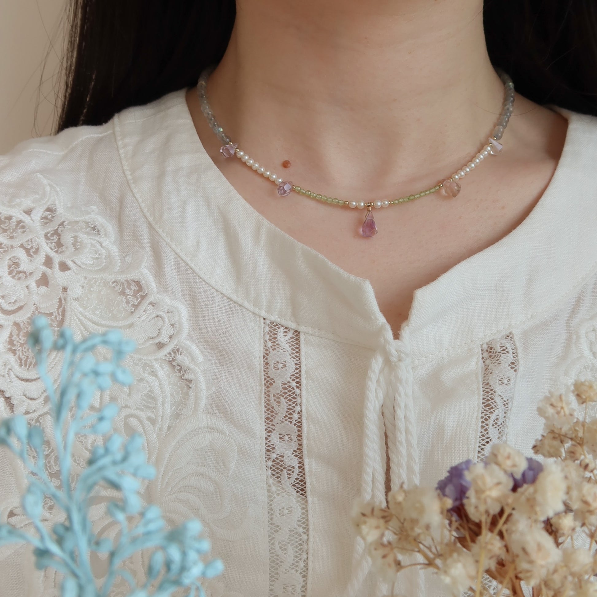 Necklace with moonstone, freshwater pearls and ametrine stones worn by a person in a white lace top, surrounded by dried flowers and a blue plant.