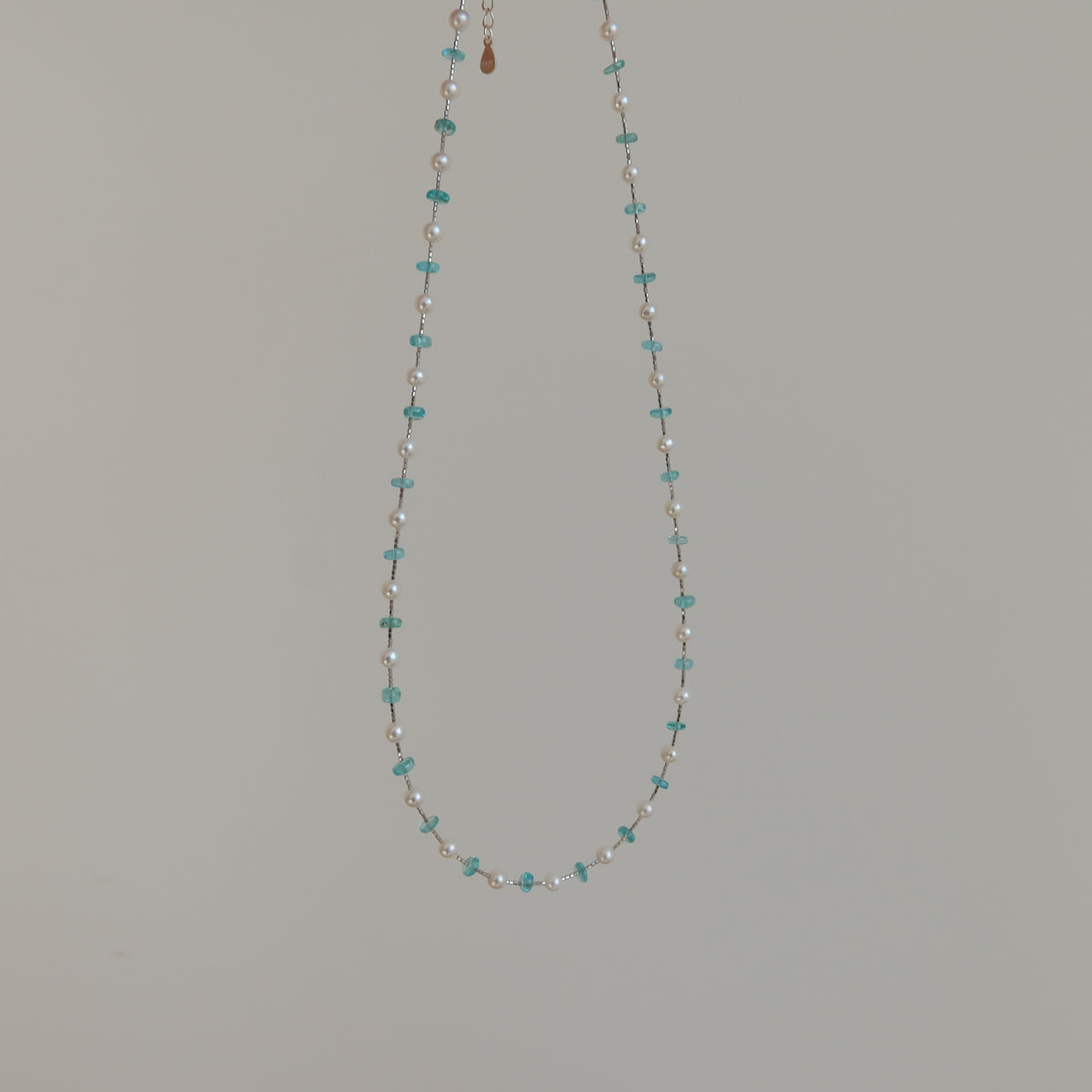 A delicate beaded necklace with small blue apatite and white pearl beads, suspended against a plain light gray background.