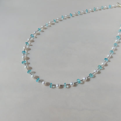 A delicate beaded necklace featuring white freshwater pearls and light-blue apatite crystals, arranged in a gentle curve against a neutral light-gray background.