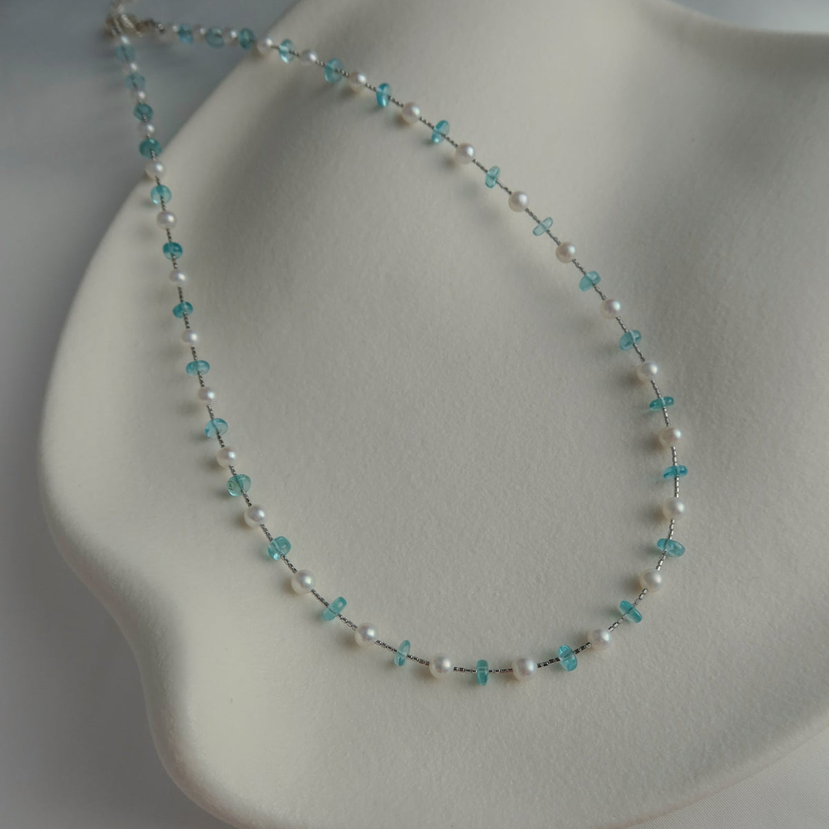 A delicate beaded necklace with white pearls and blue apatite crystal beads, displayed on a white wavy jewelry stand.