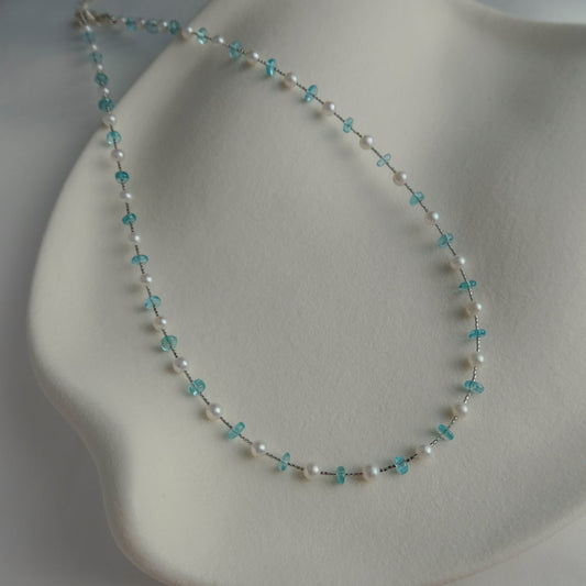 A delicate beaded necklace with white pearls and blue apatite crystal beads, displayed on a white wavy jewelry stand.