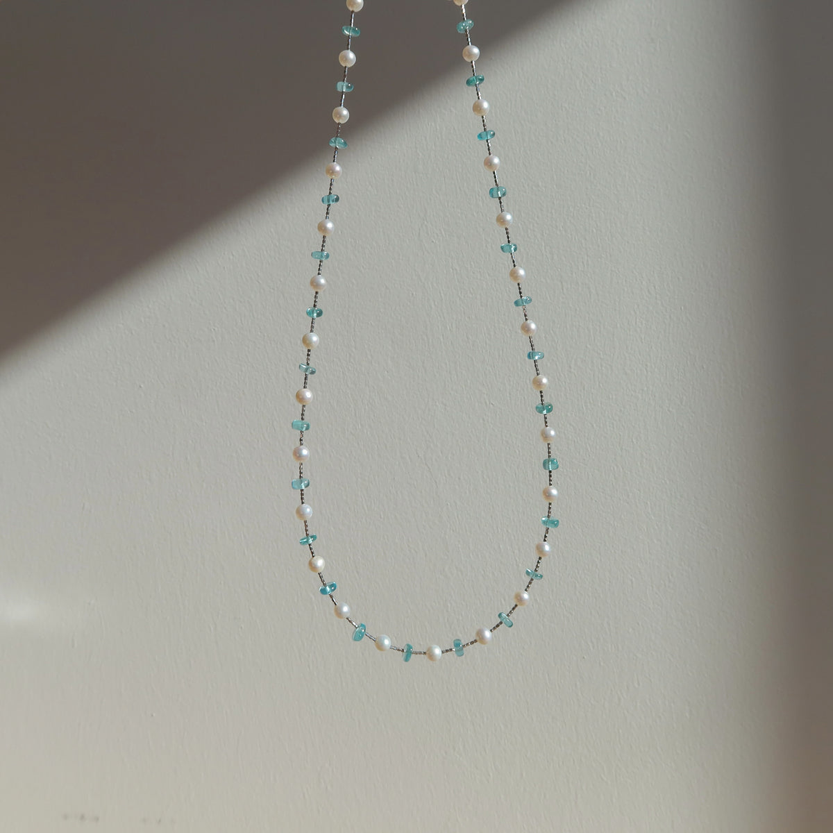 A delicate beaded necklace with white pearl beads and small blue apatited beads, hanging against a plain light gray wall.