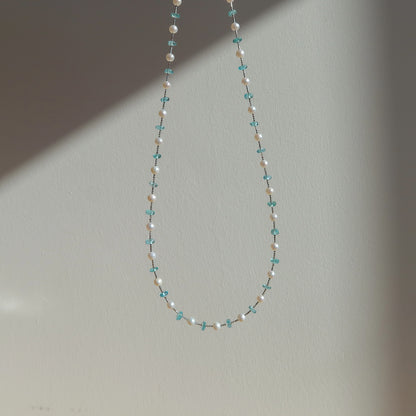 A delicate beaded necklace with white pearl beads and small blue apatited beads, hanging against a plain light gray wall.