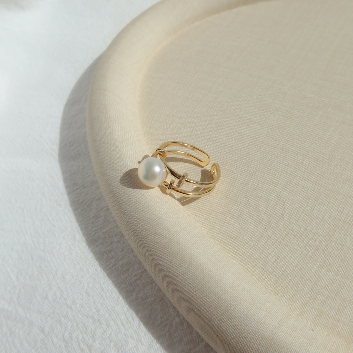 A gold-toned adjustable ring with a single white pearl, resting on a textured beige fabric surface.