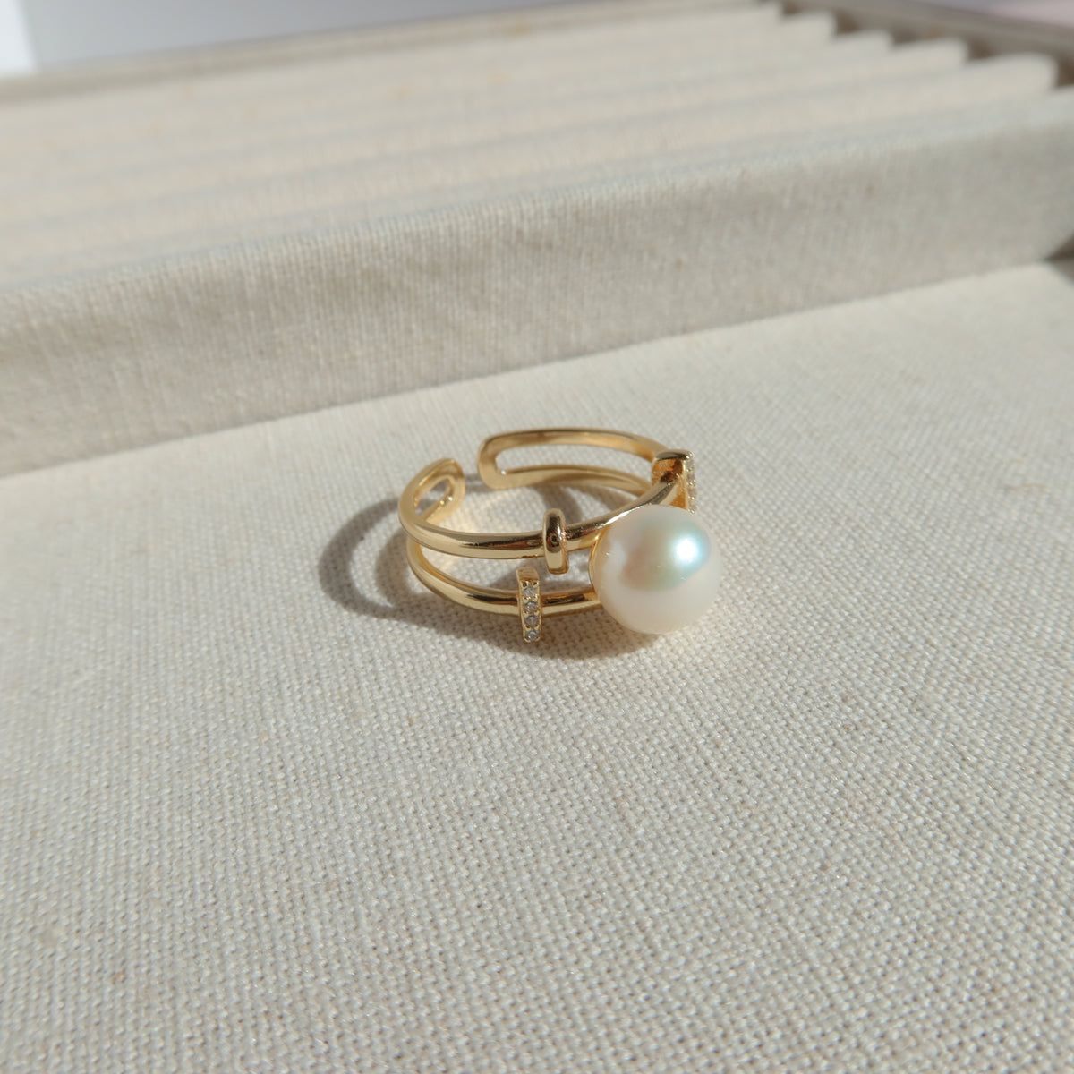 A gold-plated open ring with a single white pearl and a small clear crystal accent, displayed on a textured cream-colored jewelry box.
