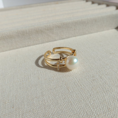 A gold-plated open ring with a single white pearl and a small clear crystal accent, displayed on a textured cream-colored jewelry box.