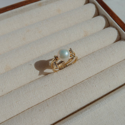 A gold ring with a single white pearl, displayed on the cream-colored padded slots of a jewelry box.