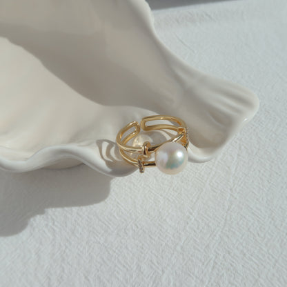 A gold-toned ring with a single round white pearl, displayed on a white ceramic shell-shaped stand against a textured white background.