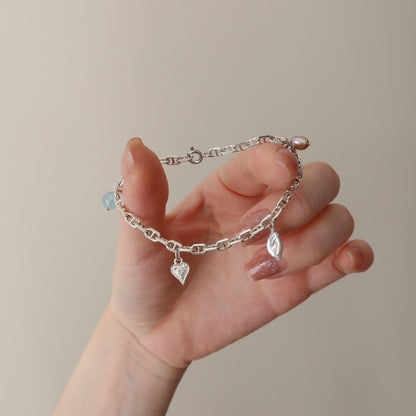 Silver chain bracelet with heart and pearl charms held in a hand against a beige background