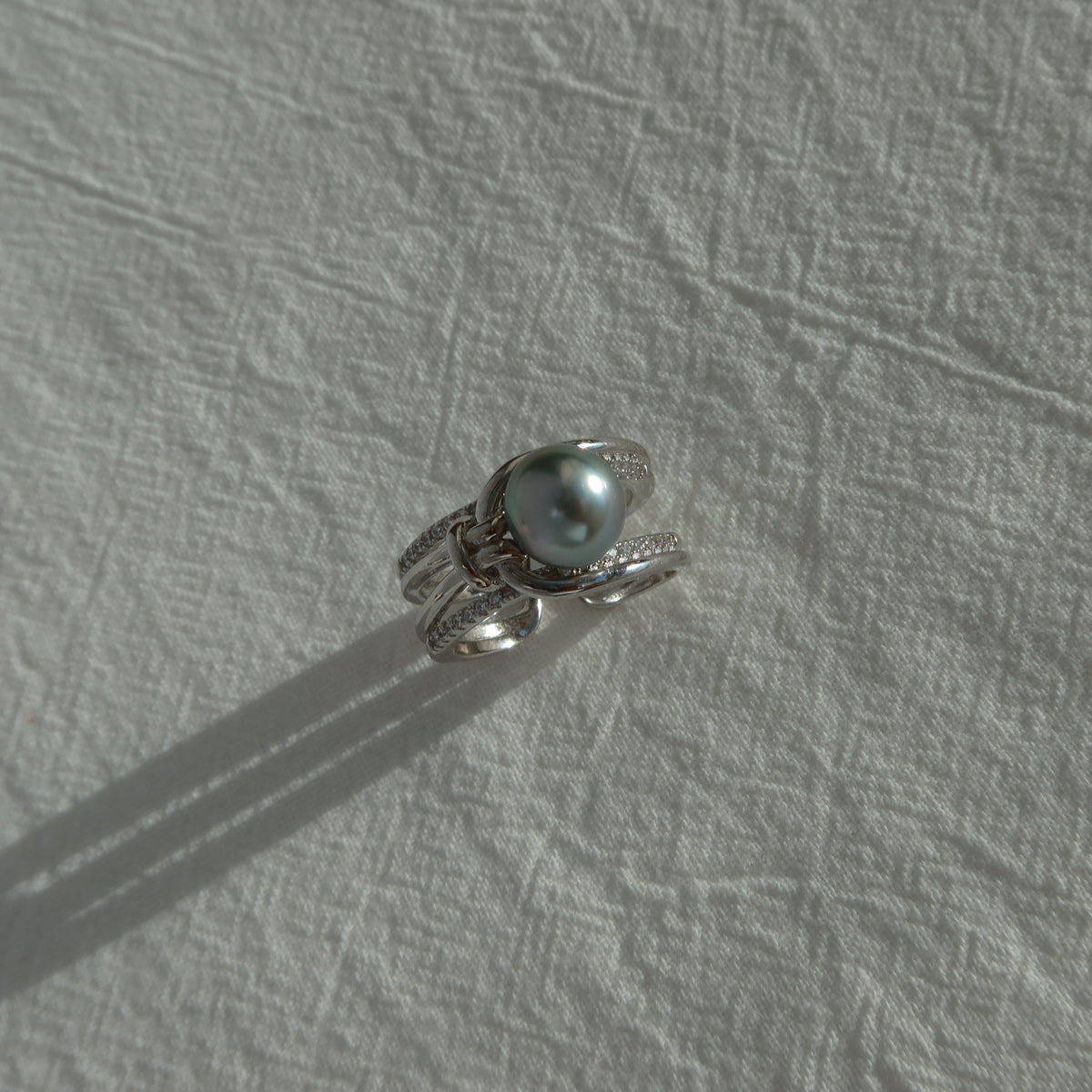 Sirene Tahitian Pearl Ring resting on a textured white surface in natural sunlight, highlighting its 8.7mm platinum-gray Tahitian pearl and twisted pavé silver band.