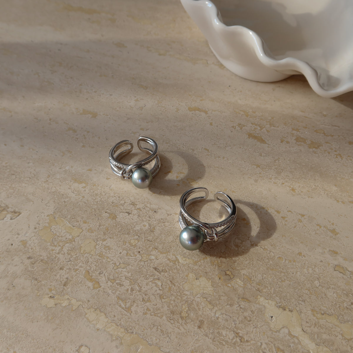 Two Sirene Tahitian Pearl Rings resting on a textured surface with a white ceramic shell dish, highlighting their 8.7mm platinum-gray Tahitian pearls and twisted pavé silver bands.