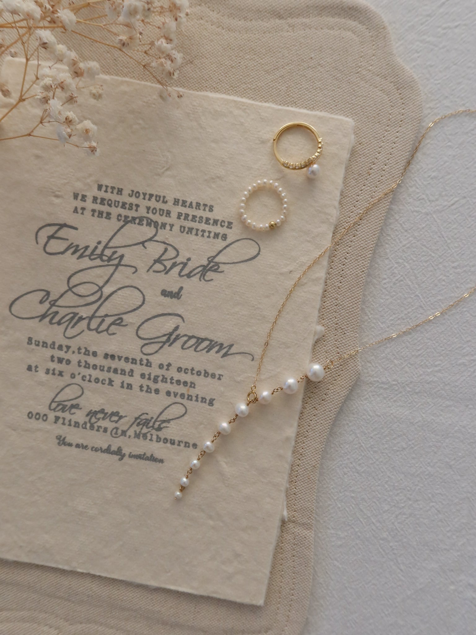 A styled flat-lay of a cream wedding invitation card with elegant black script text, accompanied by a matching cream envelope, delicate gold rings, and a pearl-and-gold necklace on a textured light background.