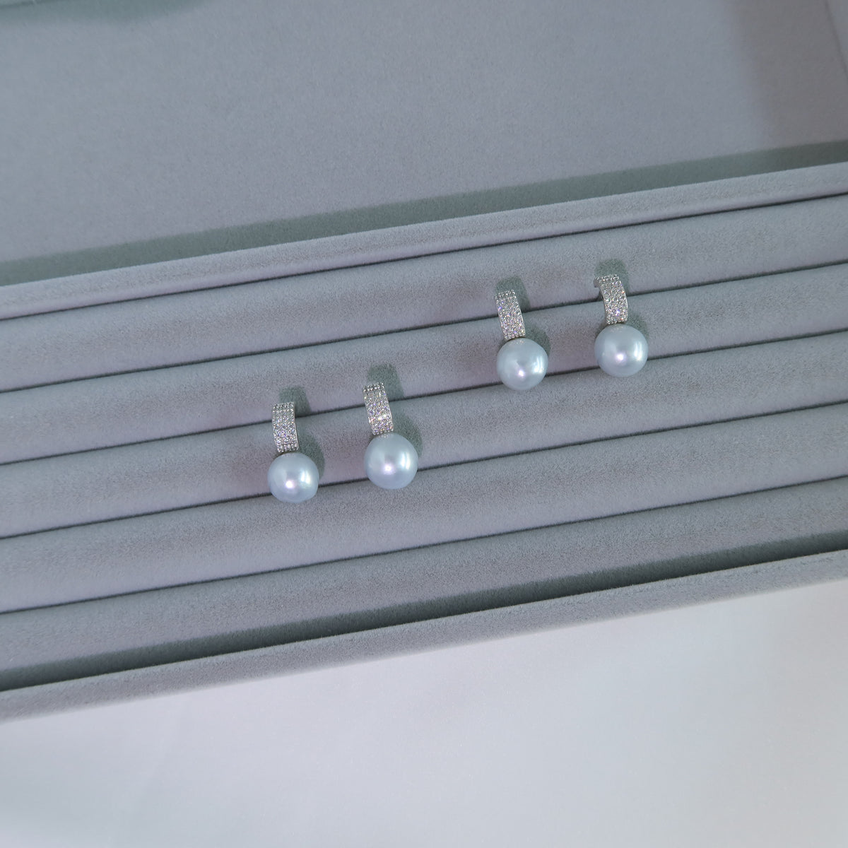 Two pairs of Nova South Sea Baroque Pearl Earrings resting on a grey velvet jewelry tray, showcasing their blue-toned white pearls and pavé huggie hoops.